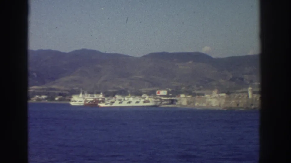 Shifted ship's-eye view of the same Greek harbor, 1976 — white ferry, stone fortress, and bare arid hills visible across the water.