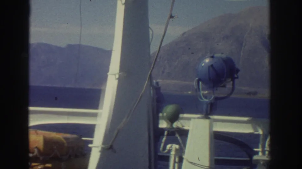 Ship deck fittings in foreground — white mast poles, blue searchlight, green deck fixture — with a dramatic bare rocky headland beyond, Greece 1976.