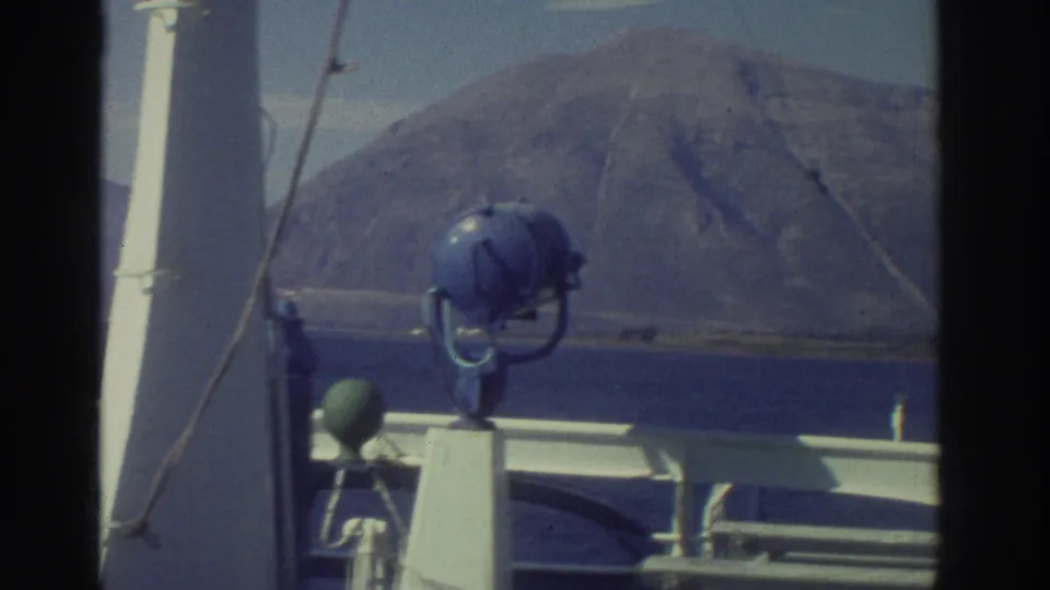 Continued ship-deck view, Greece 1976 — white masts and blue searchlight in foreground, the same dramatic rocky headland behind.