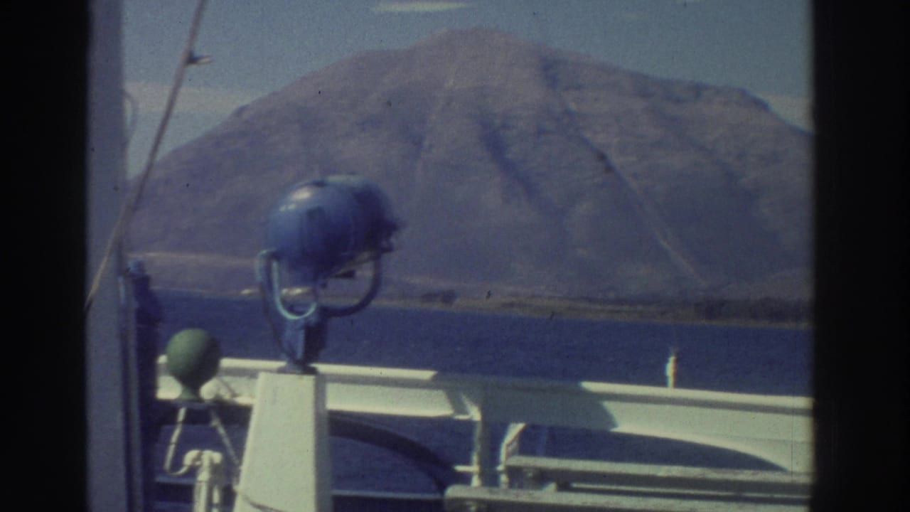 8mm film frame from aboard a ship in Greece, 1976, with a blue deck-mounted searchlight and a barren hillside visible beyond the white ship railing.