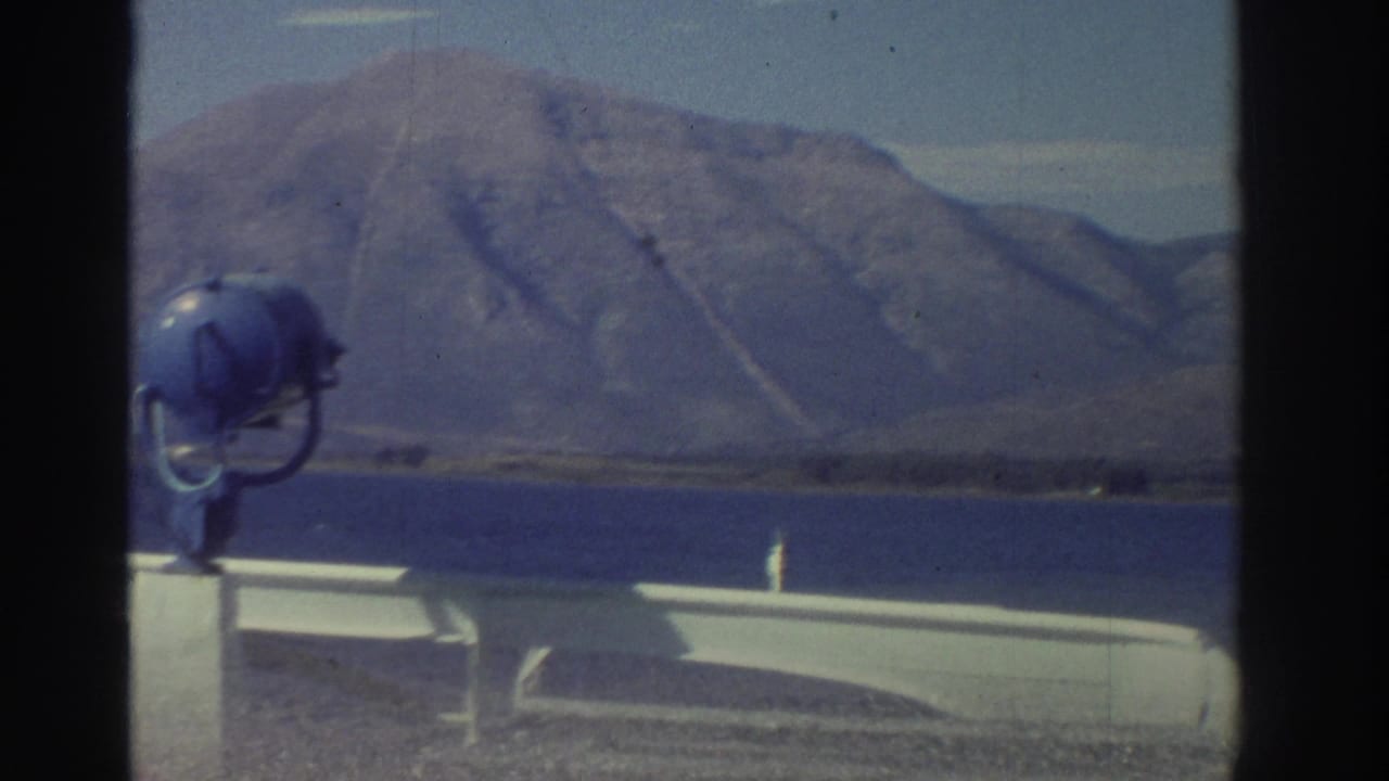 Ship's deck with a prominent blue searchlight, barren hillside and pale blue sky in the background.