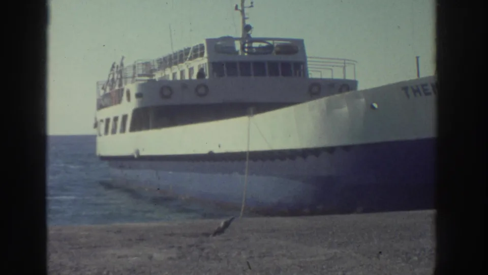 The ferry Themistoclis at a Greek pier in 1976, white hull and passenger decks visible with lifebuoys against a barren hillside backdrop.