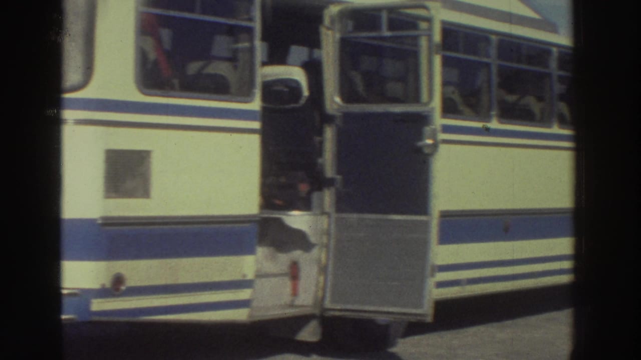 Side of a cream-and-blue Magirus Deutz coach with open door and seated passengers visible through windows, filmed in Greece circa 1976 on 8mm.