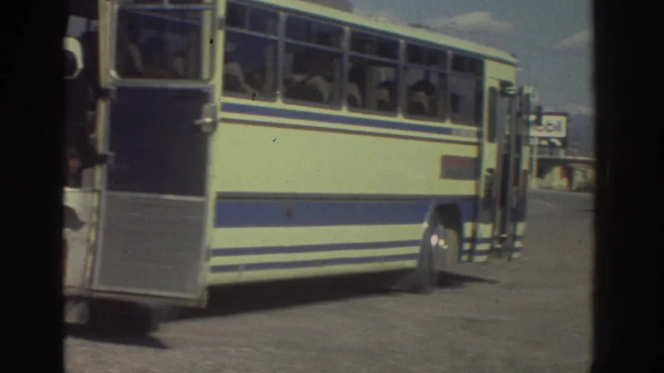 Full side of a cream-and-blue Magirus Deutz tourist coach at a Greek roadside stop circa 1976, with a Mobil branded sign and open sky visible in the background.