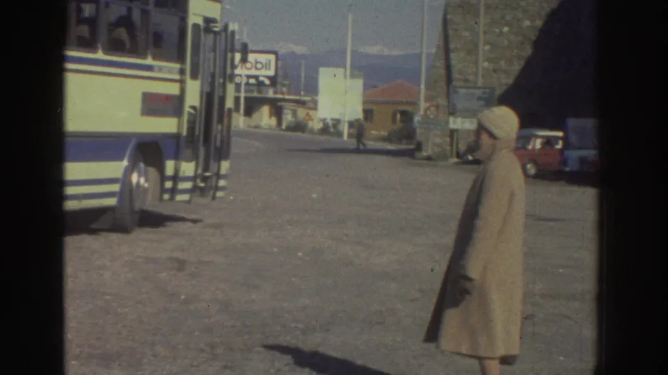 Wide view of a Magirus Deutz coach at a Greek roadside stop circa 1976, a woman in a long beige coat standing to the right, a Mobil station and low buildings visible in the mid-ground.