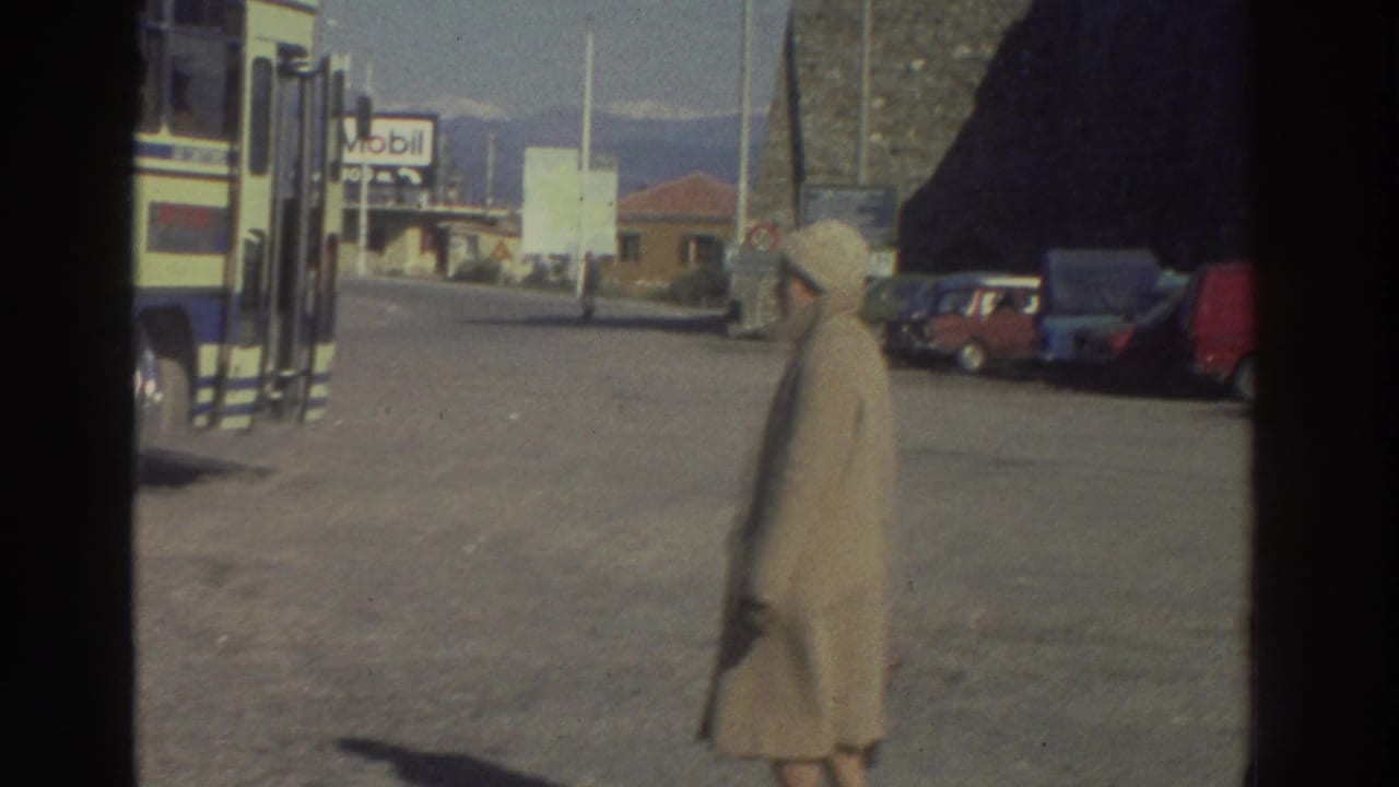 Woman in beige coat and hat crossing a roadside gravel lot at a Greek bus stop, circa 1976; Mobil station sign and stone building behind her.