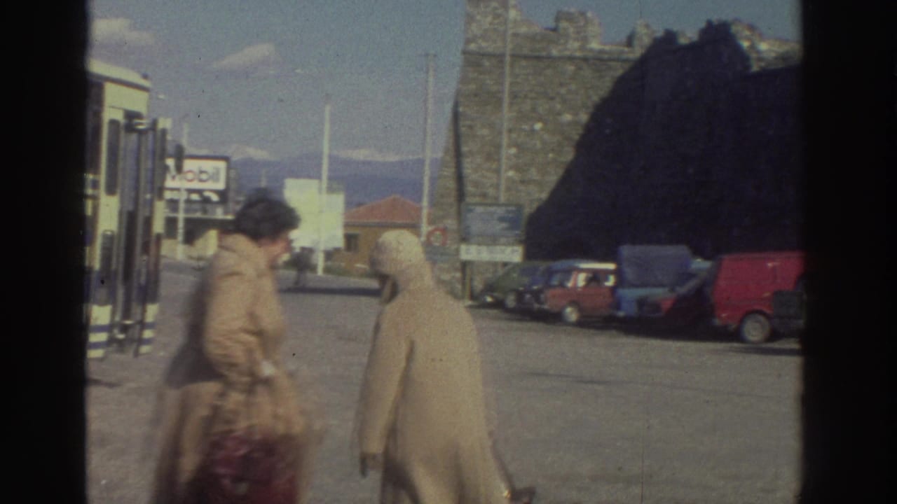Two women in beige coats moving through a Greek roadside bus stop lot, circa 1976; Mobil sign, blue and red parked cars, and mountain range visible.