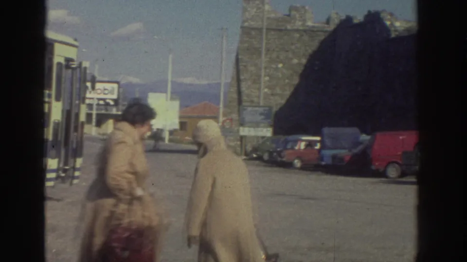 Two women in beige coats moving through a Greek roadside bus stop lot, circa 1976; Mobil sign, blue and red parked cars, and mountain range visible.