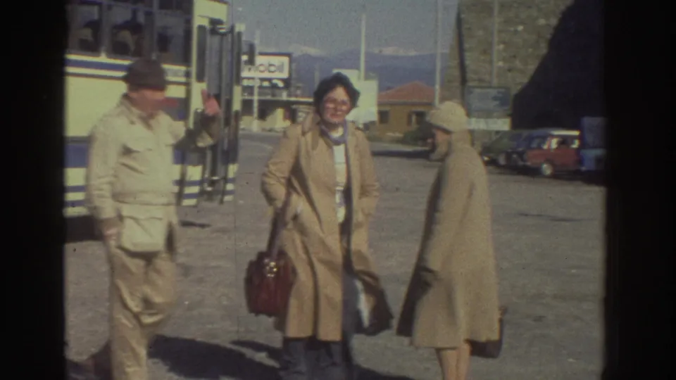 Man in uniform and two women at a Greek roadside bus stop, circa 1976; woman in beige coat and glasses smiles toward camera, red handbag in hand.