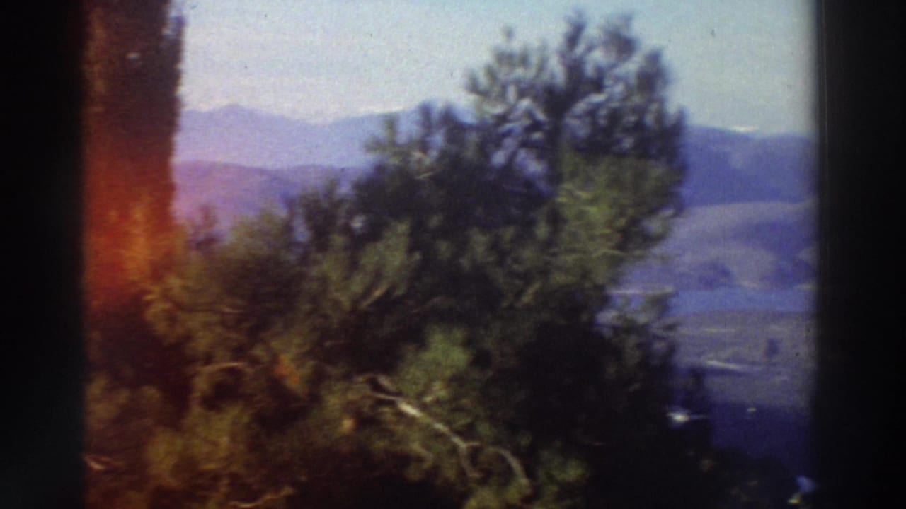 Super 8 home movie frame from Greece (c.1976) showing rolling mountain hills and a tree-framed valley under a hazy sky, with a film-edge light leak at left.