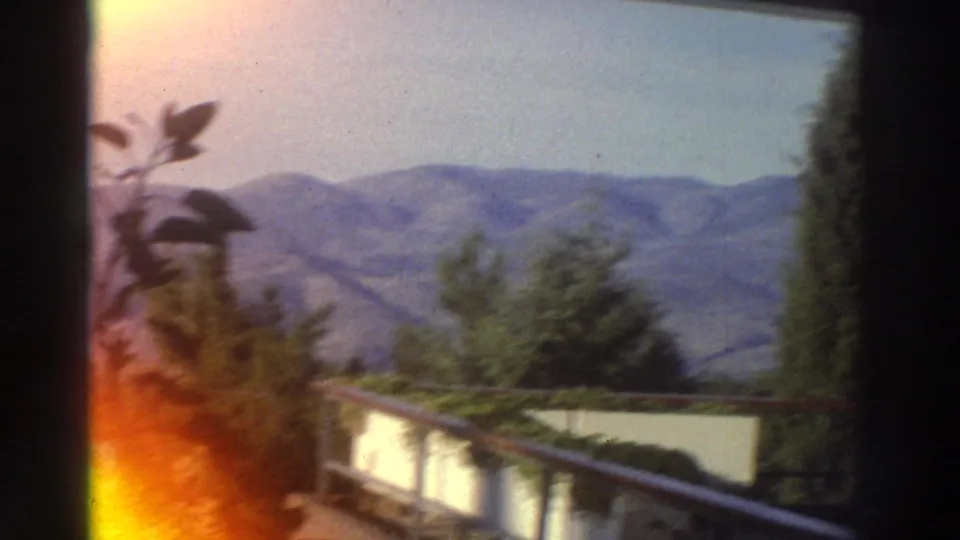 Super 8 home movie frame from Greece (c.1976) as the panning camera reveals a residential balcony railing against a mountain-hill backdrop.