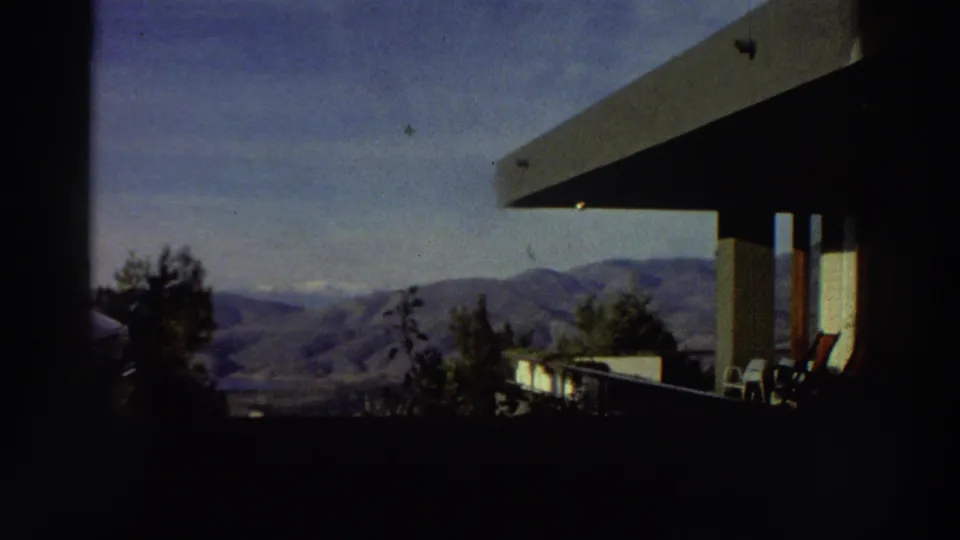 Super 8 home movie frame from Greece (c.1976) showing a residential terrace with outdoor chairs and a flat-roofed overhang, backed by mountain hills and blue sky.