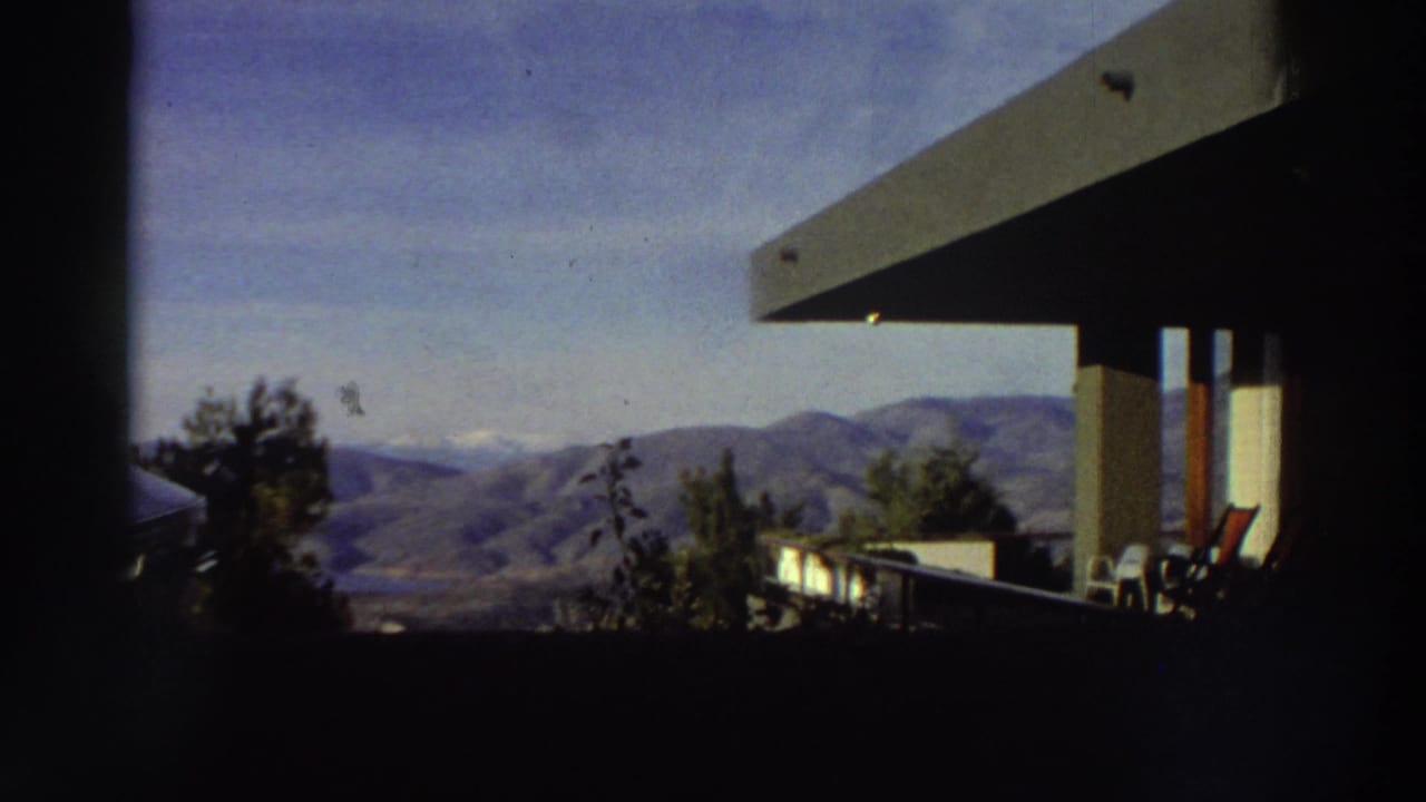 8mm home-movie shot from a modernist terrace in Greece, circa 1976, looking out over a rolling mountain range under a blue sky.