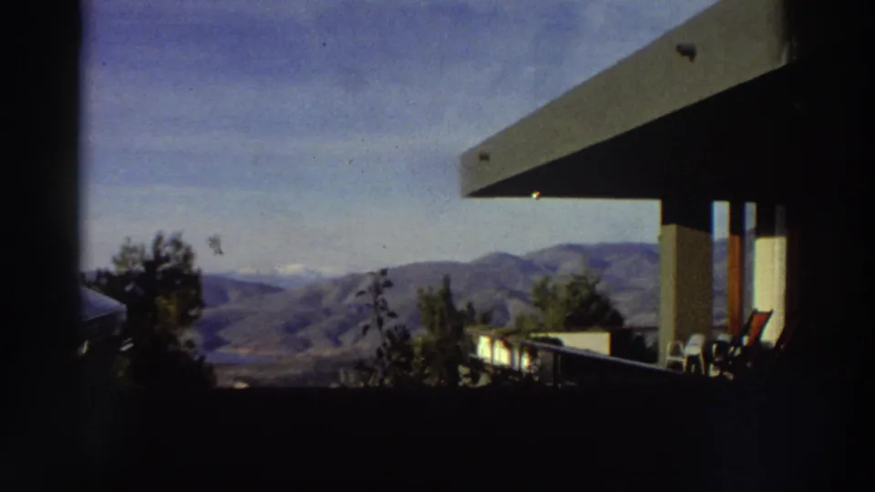 8mm home-movie shot from a modernist terrace in Greece, circa 1976, looking out over a rolling mountain range under a blue sky.