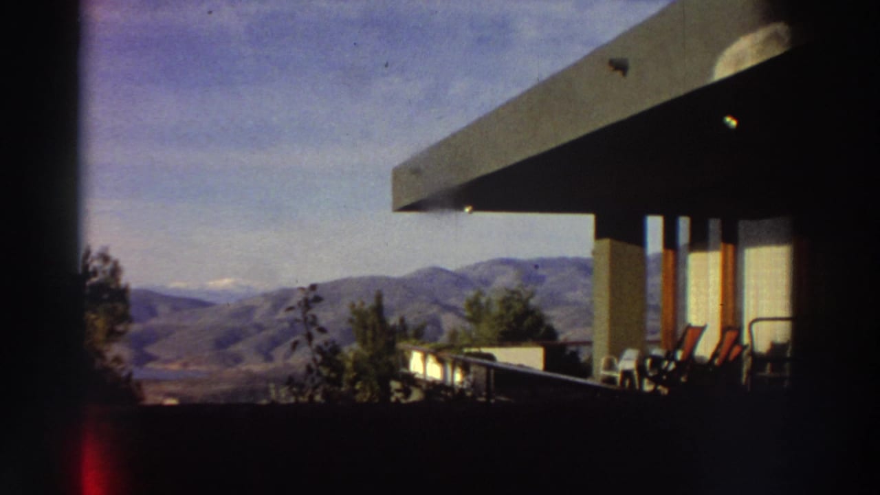 Second 8mm frame from the same modernist terrace in Greece, circa 1976, showing the mountain panorama with terrace furniture under open sky.