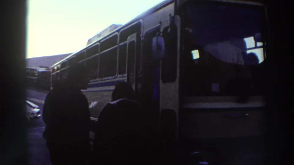 8mm home-movie frame of a dark touring coach with side windows, silhouetted passengers in the foreground, filmed in Greece circa 1976.
