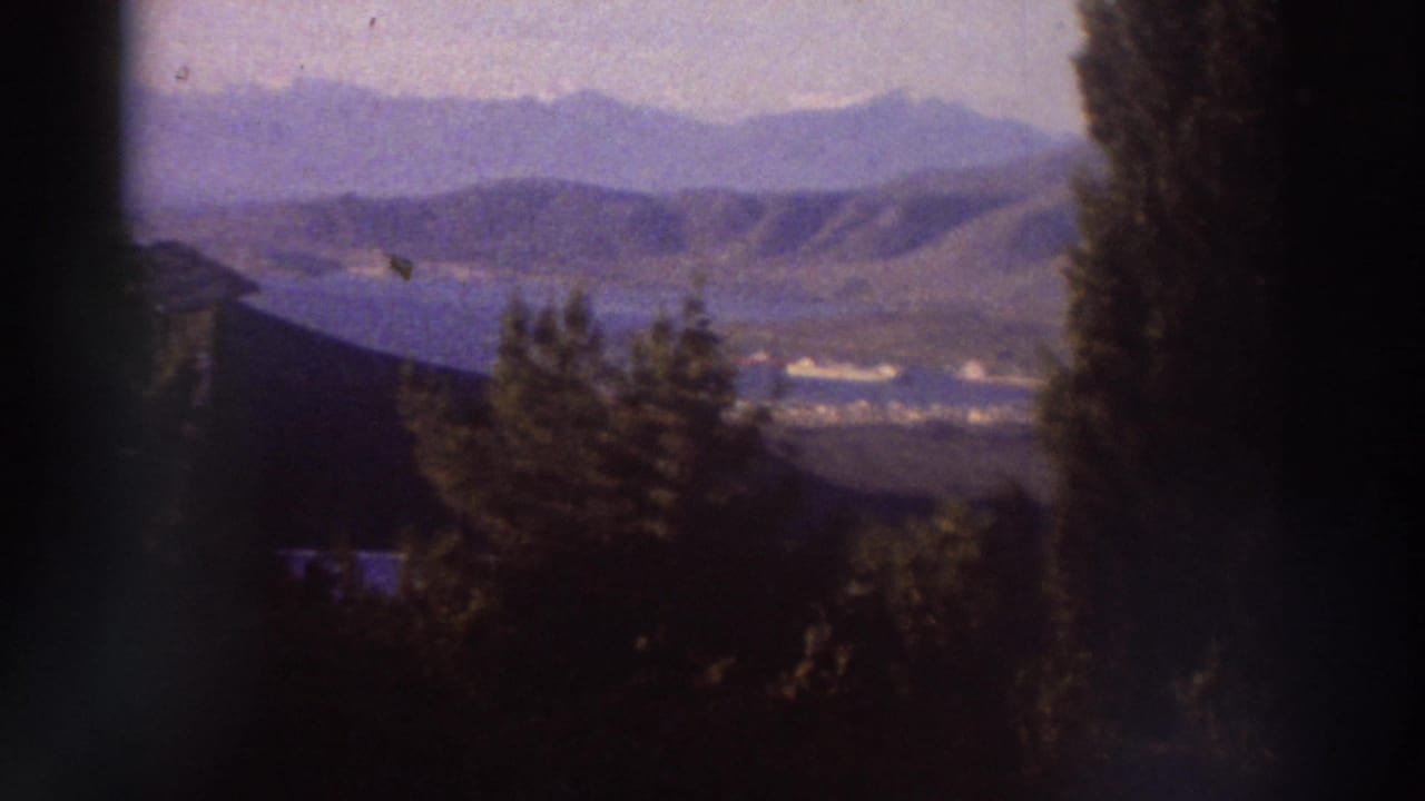 Wide panoramic view of layered mountain ridges and a valley lake or coastal inlet in Greece, circa 1976, framed by cypress trees with characteristic 8mm haze.
