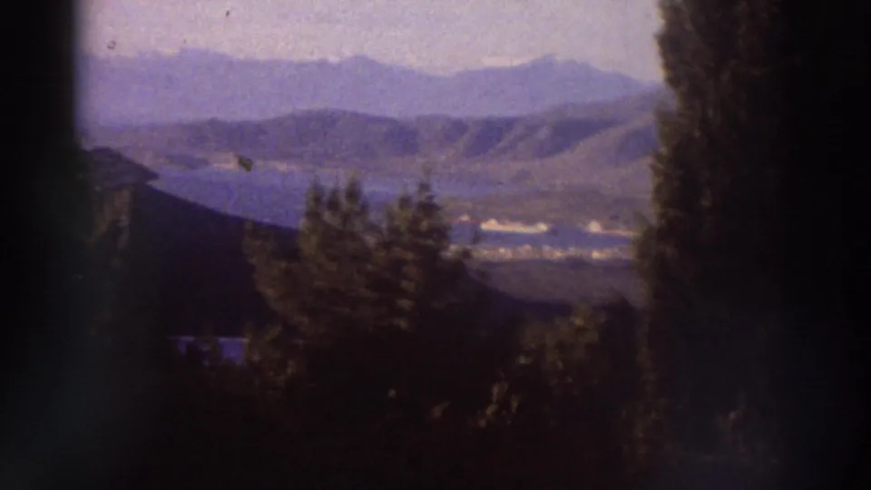 Wide panoramic view of layered mountain ridges and a valley lake or coastal inlet in Greece, circa 1976, framed by cypress trees with characteristic 8mm haze.