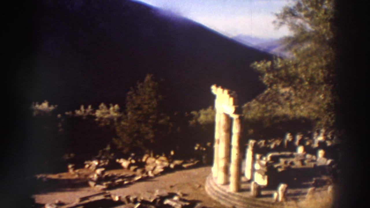 Ancient Greek columns and ruins with camera panned slightly right, scattered stone blocks across site, trees and mountains behind, warm 8mm tones.