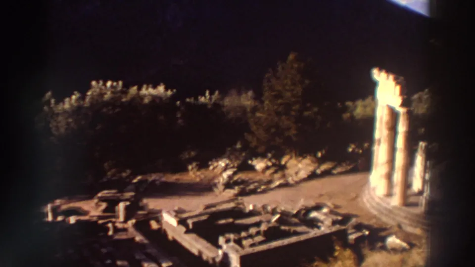 Three tall columns on a circular stepped base dominate the frame at a Greek ruin site, 1976, with strong backlighting and visible 8mm grain.