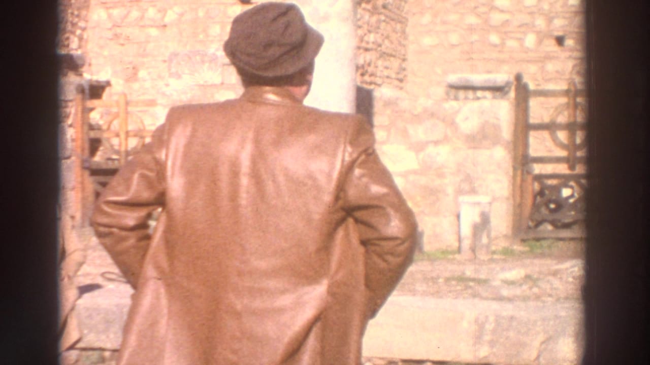 Man in long brown leather coat and rounded dark hat faces ancient stone ruins from behind; 8mm home movie, Greece, 1976.