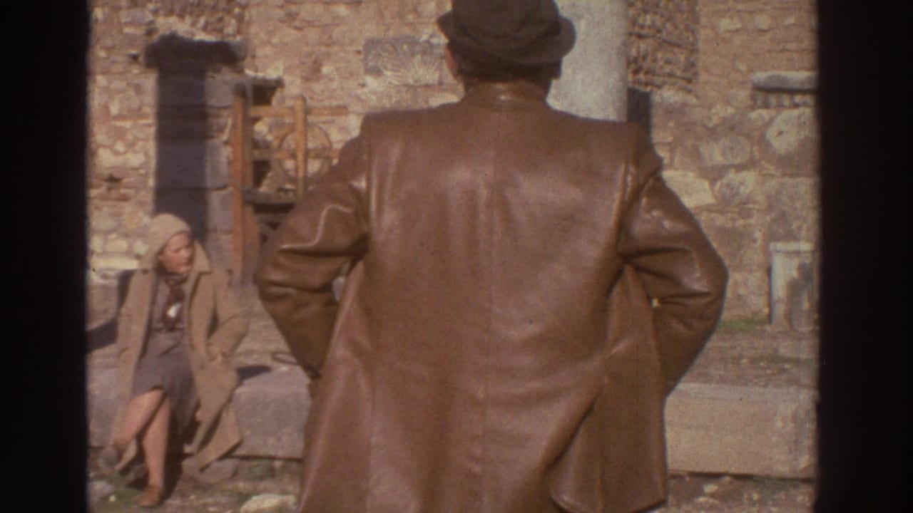 Man in brown leather coat stands right of frame while a woman in a light coat sits on stone ruins to the left.