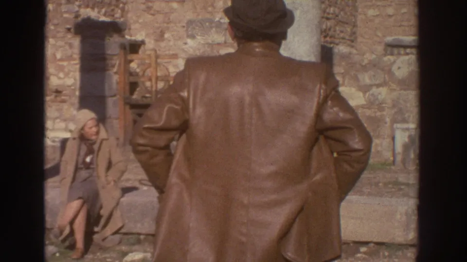 Man from behind and a seated woman in a belted light coat rest amid ancient stone ruins; 8mm home movie, Greece, 1976.