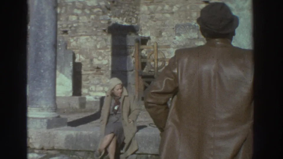 Wide shot of man and seated woman amid ancient stone columns and rubble at a Greek archaeological site; 8mm home movie, 1976.