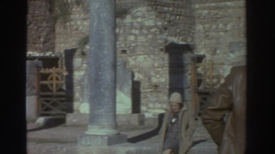 Tall cylindrical ancient stone columns at a Greek archaeological site with two visitors in mid-1970s dress; 8mm home movie, 1976.