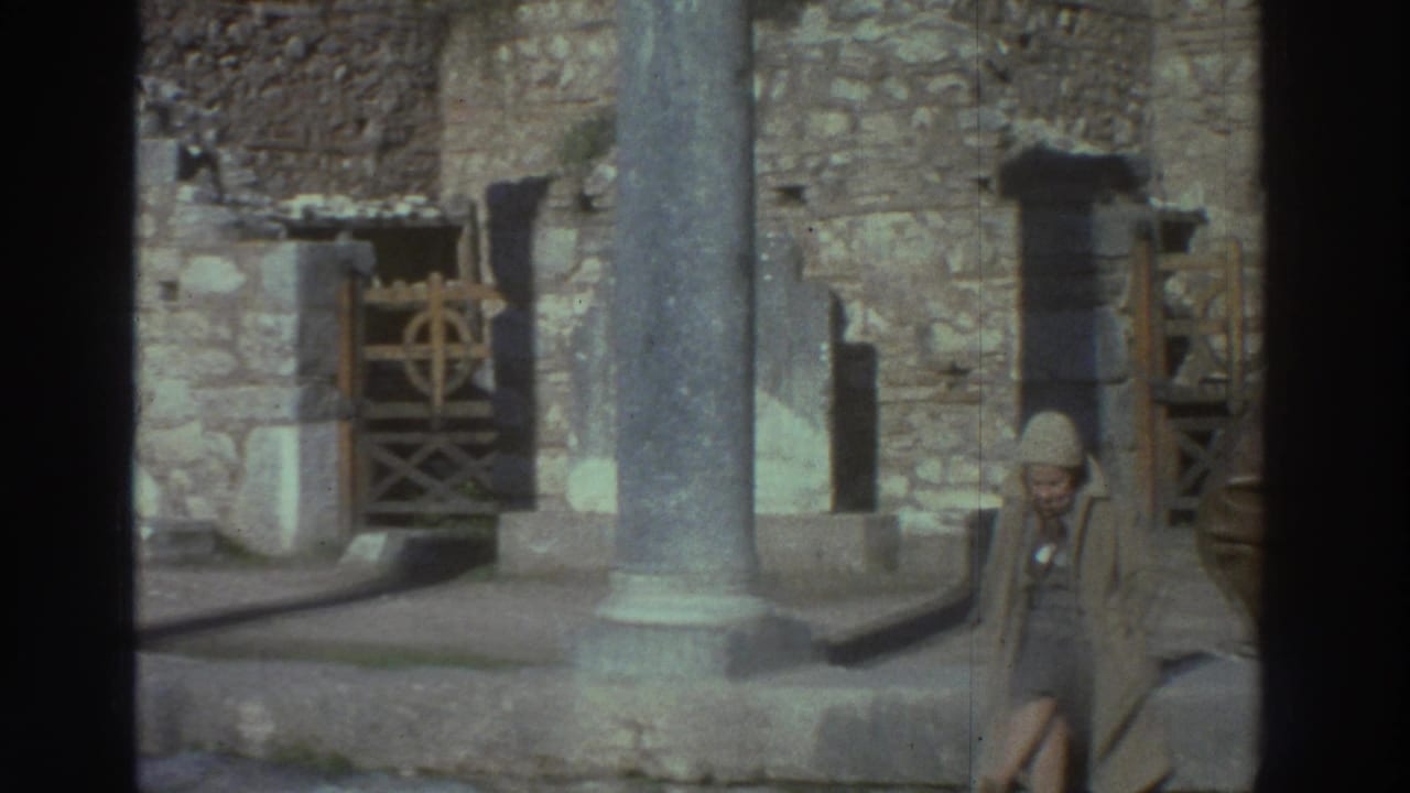 Super 8 home movie, Greece 1976: interior of ancient ruins with stone columns, ashlar walls, a wooden gate with decorative ironwork, and an adult visitor in 1970s travel attire.