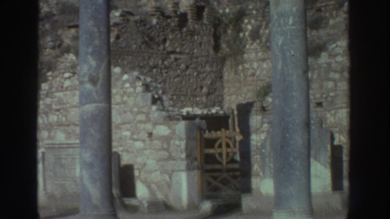 Ancient Greek ruins with stone columns and crumbling masonry walls; the wooden gate with circular ironwork remains visible in the background after the visitor has moved out of frame.
