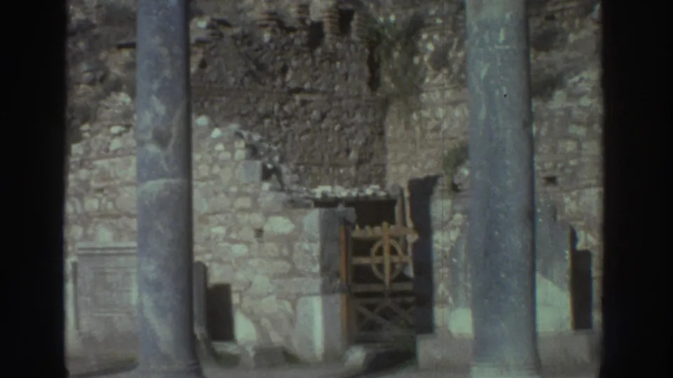 Super 8 frame, Greece 1976: stone columns and crumbling ashlar walls of ancient ruins, with the decorative wooden gate still visible as the camera begins to pan away from the visitor.