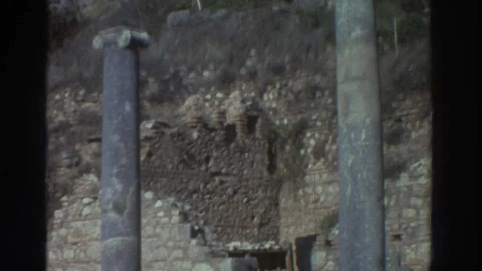Mid-pan Super 8 frame, Greece 1976: broken column stubs and a taller partial column in the foreground, stone rubble on the ground, and scrubby vegetation on a rocky hillside beyond.