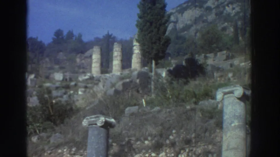 Super 8 exterior wide shot, Greece 1976: ruined columns in a landscape of cypress trees and rocky hills under a blue-shifted sky, partial column capital visible in the lower foreground.