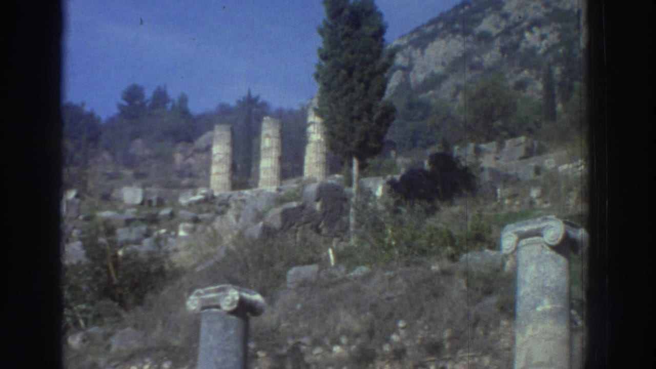 Distant ancient ruins on a sunlit hillside with a row of standing columns and a lone column capital in the foreground.