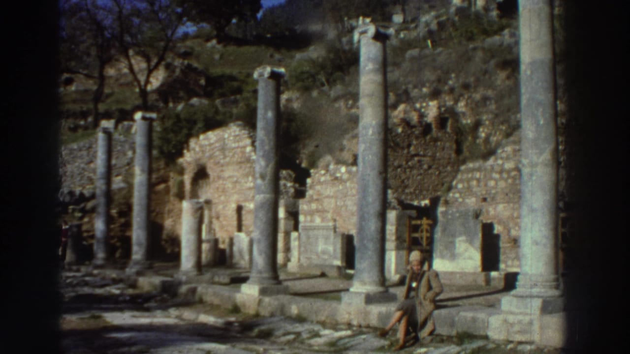Colonnaded ancient walkway with tall stone columns and a seated adult figure, ruined stone walls visible in the background.