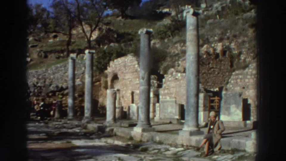 1970s 8mm footage of an ancient colonnaded street in Greece with a seated visitor and background tour group, ruined walls with arched openings behind.