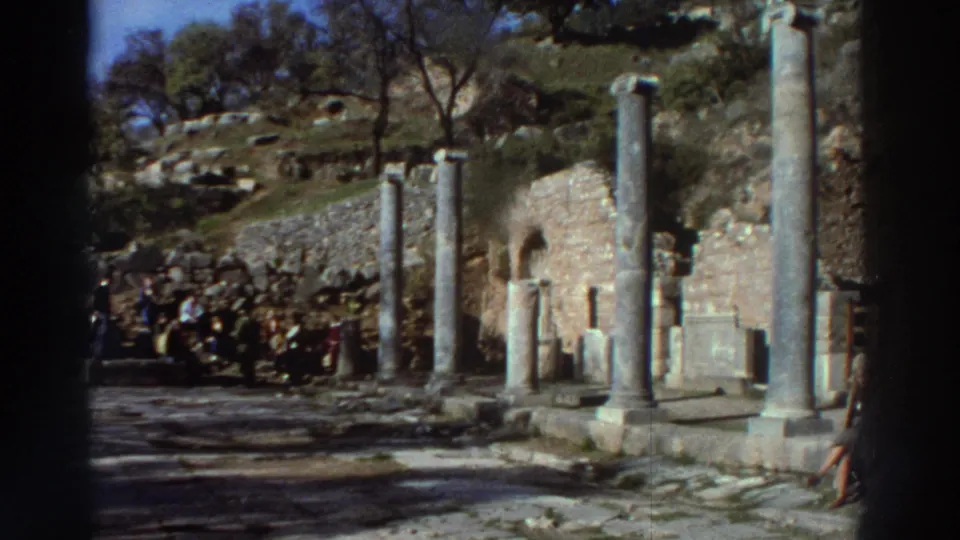 1970s 8mm home movie showing visitors gathered among ancient stone columns at a Greek archaeological site, ruined walls and hillside trees behind.
