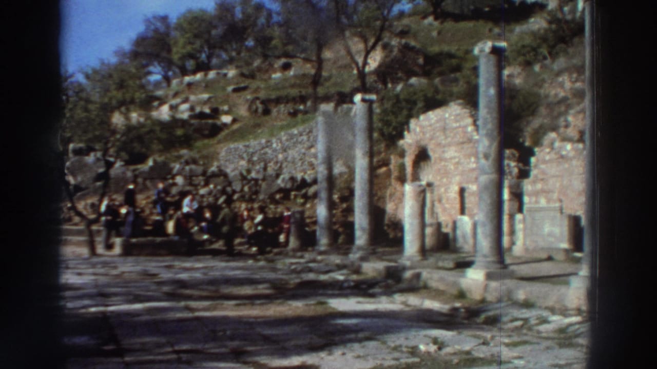 Wide shot of an ancient Greek archaeological site, circa 1976; tourists grouped at left amid standing stone pillars and ruined stone structures on a hillside.