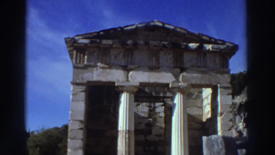 Small Doric treasury-style Greek temple structure with columns and pediment, filmed on 8mm in Greece in 1976 against a vivid blue sky.