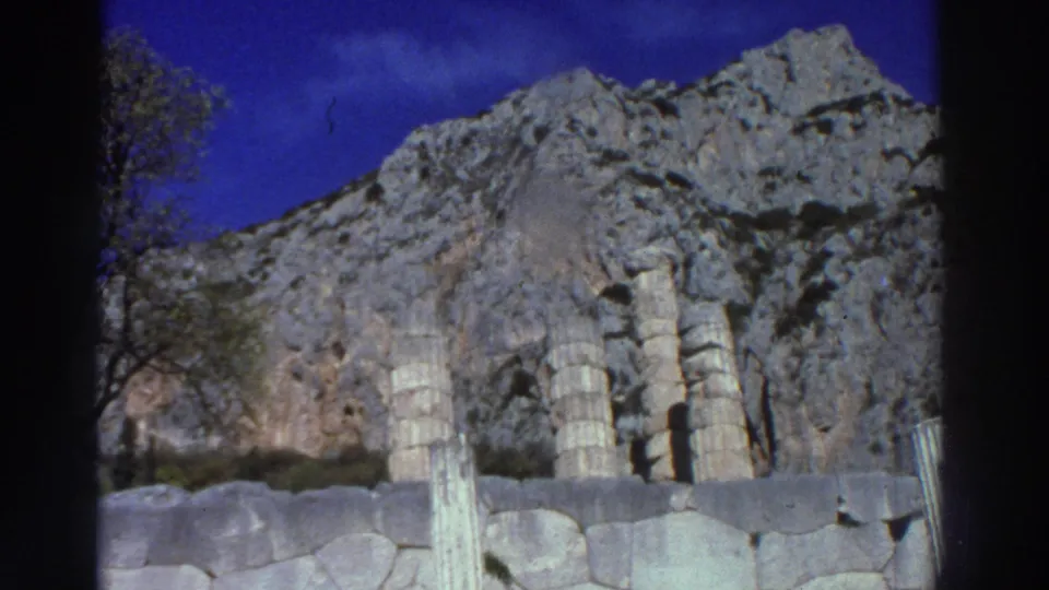 Partially preserved ancient column drums at a Greek archaeological site, with a dramatic limestone cliff in the background, filmed on 8mm circa 1976.