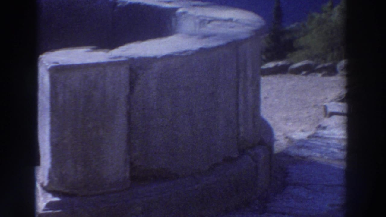 Large ancient stone column drums and block foundations on a paved terrace, deep blue sky above, green trees in background.