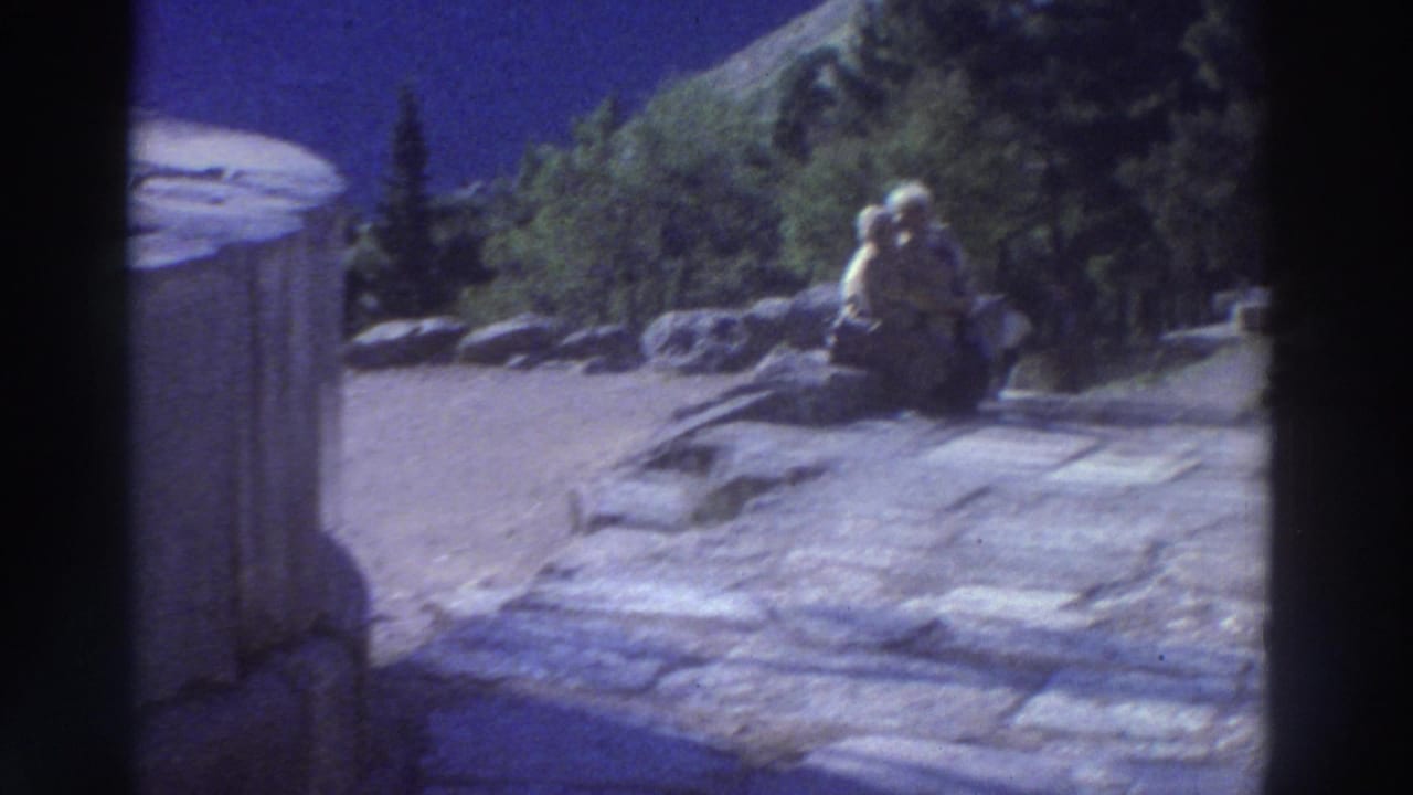 Wide view of an ancient stone site with a couple seated on rocks in the mid-distance, conifer trees and rocky hillside behind, blue sky.