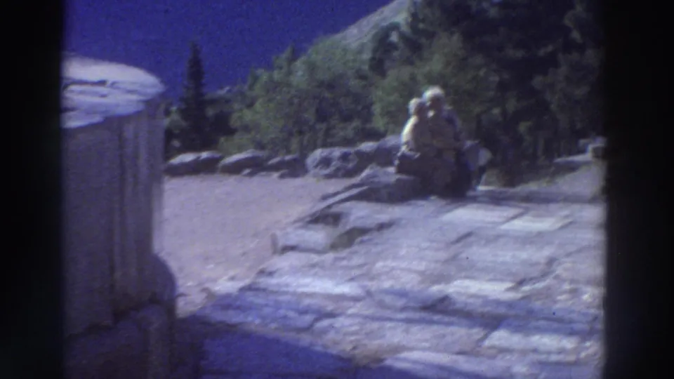 Wide shot of a couple resting on rocks amid ancient Greek ruins, conifer trees and a hillside visible behind, filmed on 8mm circa 1976.