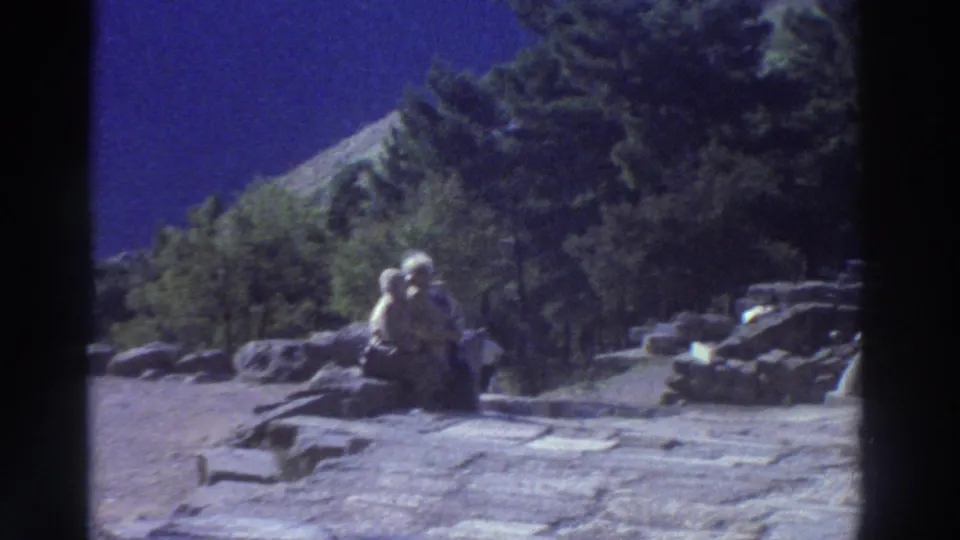 Two visitors sitting on rock rubble at a Greek archaeological site with green trees and a rocky hillside behind, filmed on 8mm circa 1976.