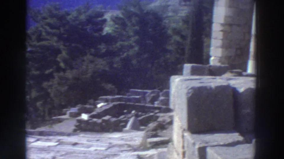 Partial ancient stone column and scattered cut stone blocks on a terrace at a Greek site, filmed on 8mm circa 1976, with conifer trees behind.