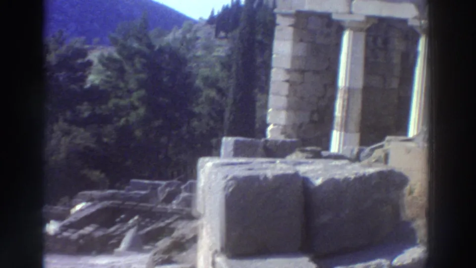 8mm home movie frame from Greece, c.1976, showing Doric stone columns and scattered marble blocks against a wooded mountainside.