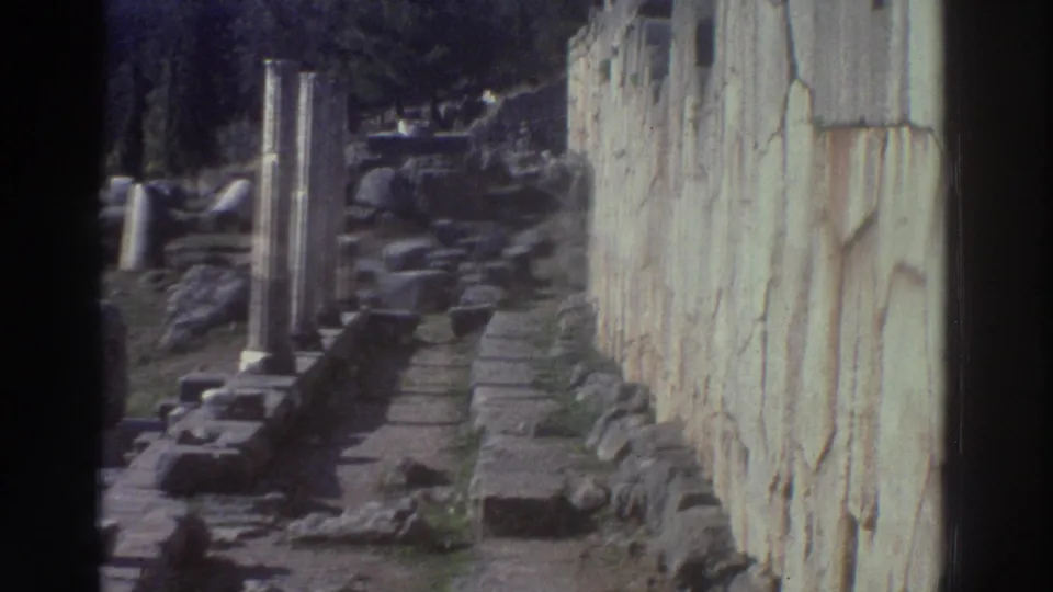 8mm home movie frame from Greece, c.1976, showing a stone-paved path through ruins flanked by column stumps and a massive ancient wall.