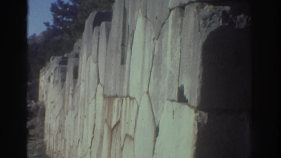 8mm frame of towering ancient stone masonry with an arched gateway opening, trees and blue sky beyond, at what appears to be an ancient Greek site, c.1976.