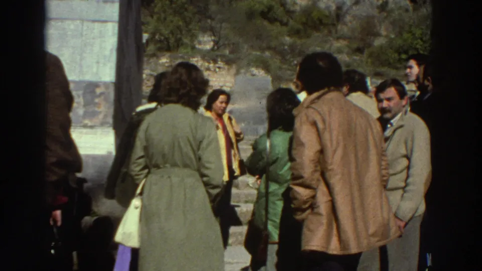 8mm home-movie frame of approximately ten adults in 1970s earth-tone coats and jackets listening at an ancient Greek archaeological site, c.1976.
